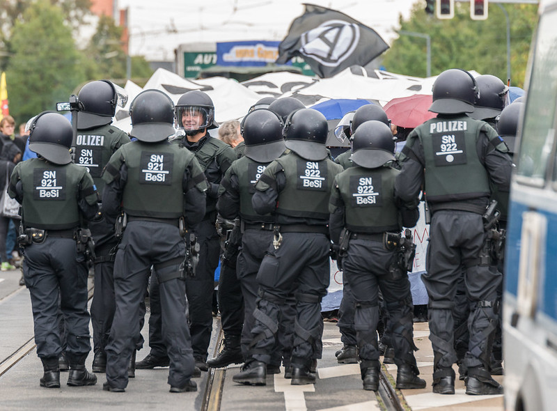 Schwach in Sepia eingefärbtes Foto von einer antifaschistischen Demonstration in Dresden, vor der sich eine Reihe behelmter Polizisten gezogen hat, die sich scheinbar teilweise gut gelaunt unterhalten.