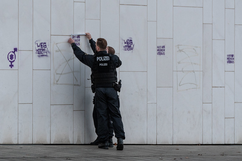 Schwach in Sepia eingefärbtes Foto zweier Polizisten, die mit einem Zollstock oder ähnlichem Werkzeug die Größe von gesprühten Stencils gegen den §219a messen.
