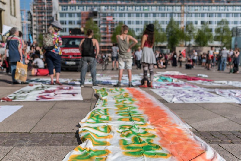 Schwach in Sepia eingefärbtes Foto einer Kundgebung auf dem Leipziger Augustusplatz. Es liegen Transparente auf dem Boden ausgebreitet und unscharf sind mehrere Personen von hinten zu sehen.